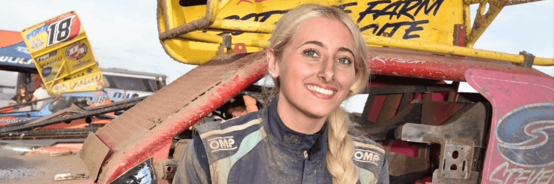 Smiling woman in racing suit holds trophy, sitting on a mud-splattered car with yellow spoiler, celebrating victory.
