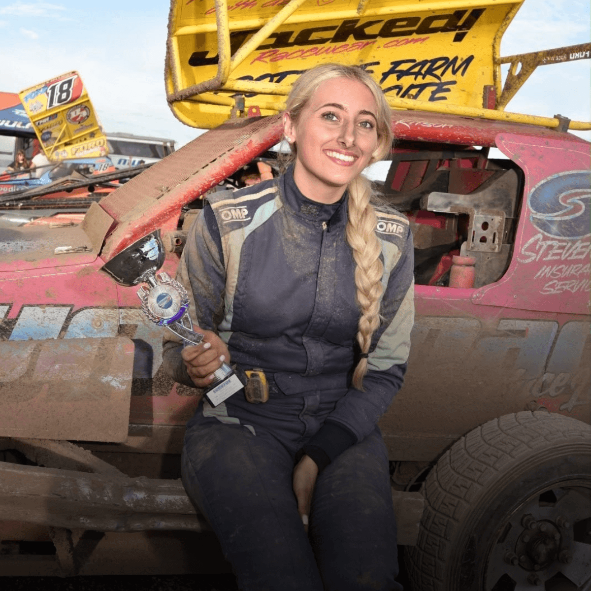 Smiling woman in racing suit holds trophy, sitting on a mud-splattered car with yellow spoiler, celebrating victory.